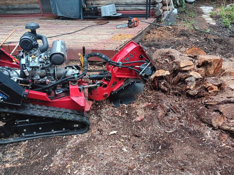 stump-grinding-cabin-jackson-hole - Teton Rope Access Close-up of stump grinding machine removing tree stump near log cabin deck in Jackson Hole, Wyoming